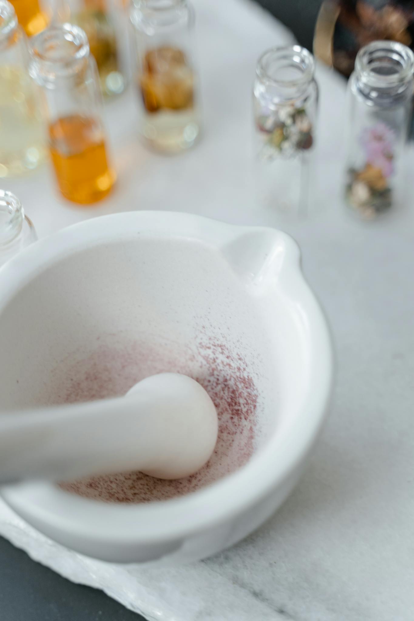A close-up of a mortar and pestle and glass bottles with herbs and oils on a marble surface.